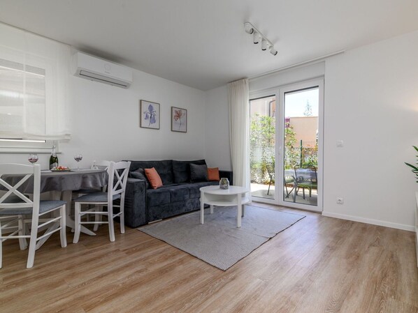 A modern living room with a dark grey sofa, white chairs, a small round table, and a glass door leading to a balcony with plants.