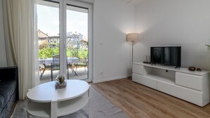 A modern living room with a white sofa, a round table with a vase, a TV on a white stand, and a sliding glass door leading to a balcony.