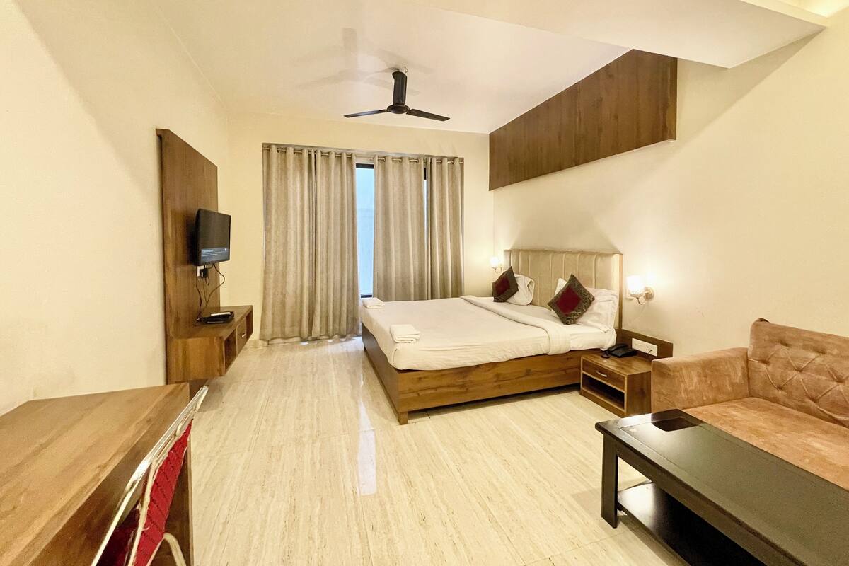 Une chambre d’hôtel comprenant un lit, une télévision, un ventilateur de plafond, une table de chevet en bois, un canapé et une fenêtre avec des rideaux.
