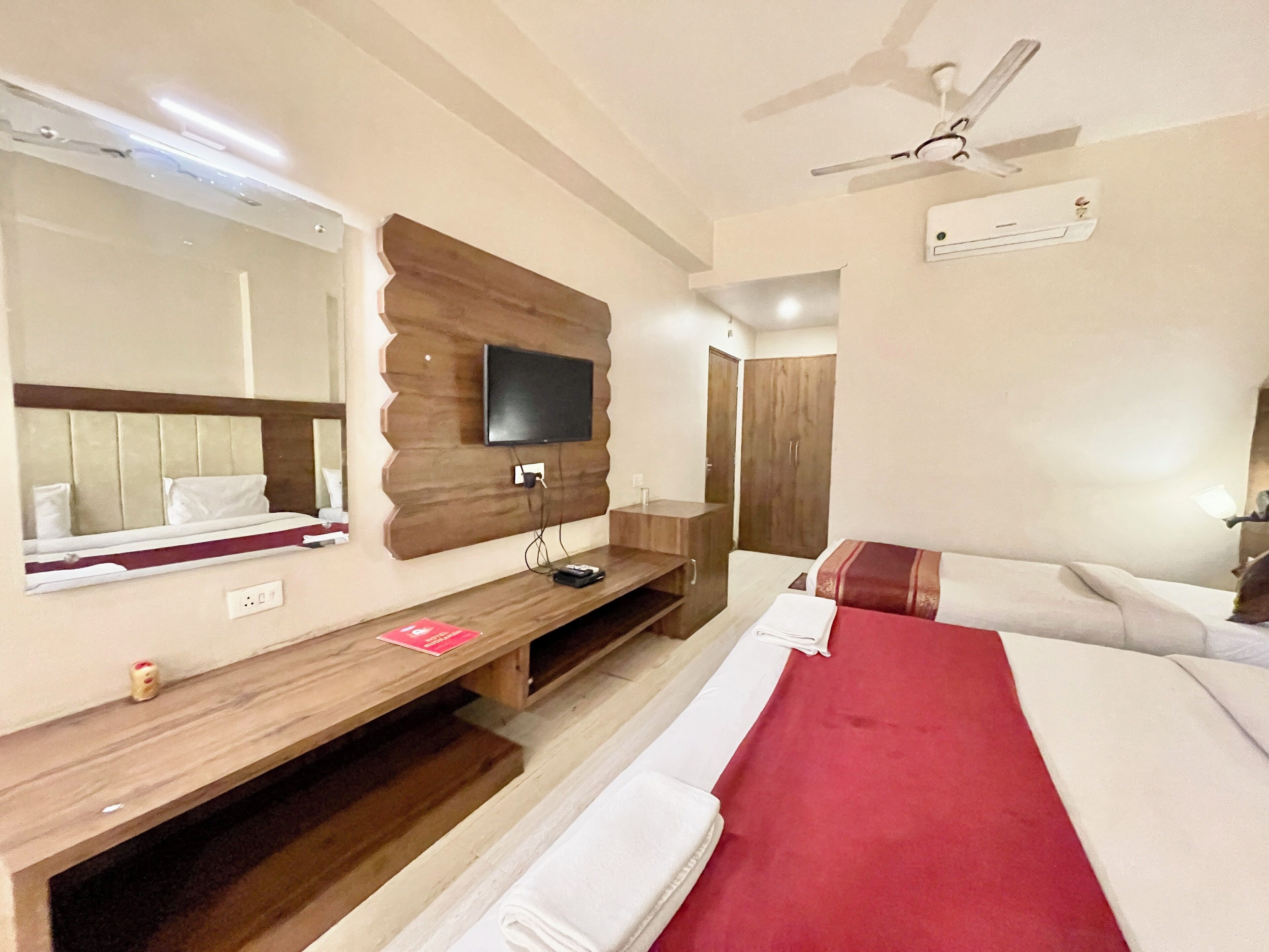 A hotel room with a bed, a TV mounted on a wooden panel, a mirror, a wooden desk, and a wardrobe.