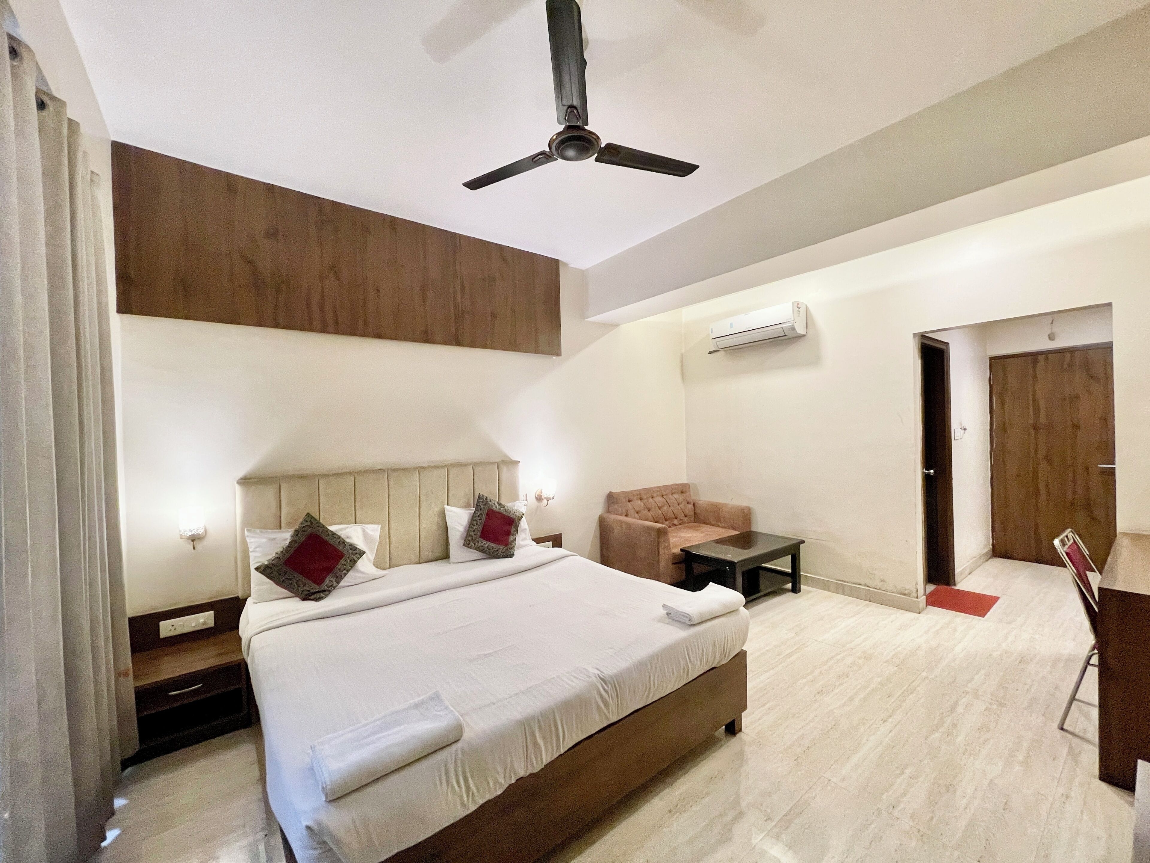 A modern hotel room with a large bed, a ceiling fan, an air conditioning unit, a small table, a chair, and a door.