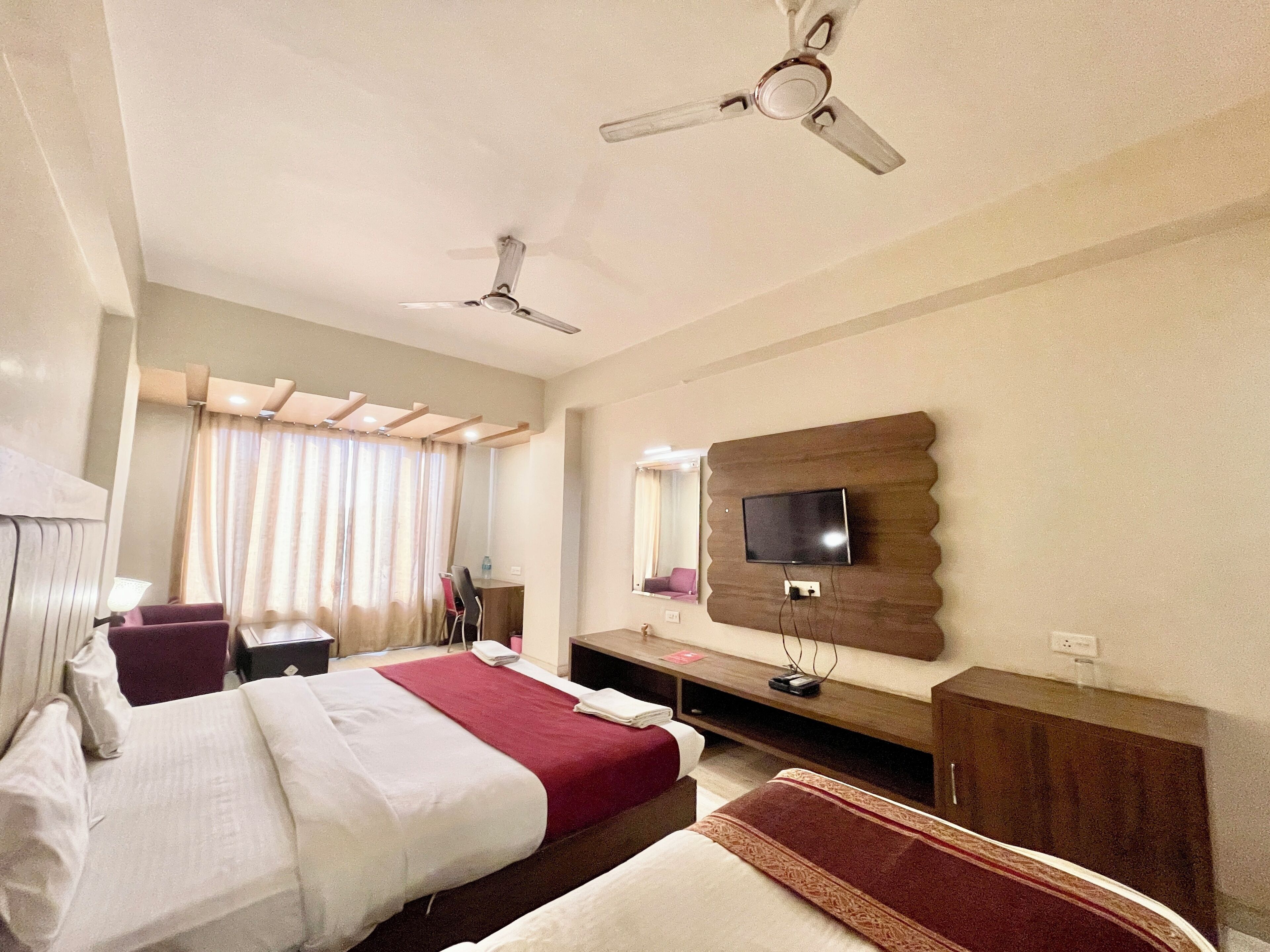 Habitación de hotel con dos camas, un televisor montado en un panel de madera, ventilador de techo y un escritorio con silla.