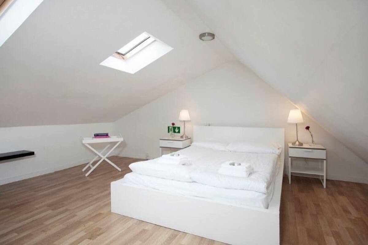Ein Schlafzimmer mit einem Bett, zwei Nachttischen, einem kleinen weißen Tisch, einer Deckenleuchte und einem Oberlicht.