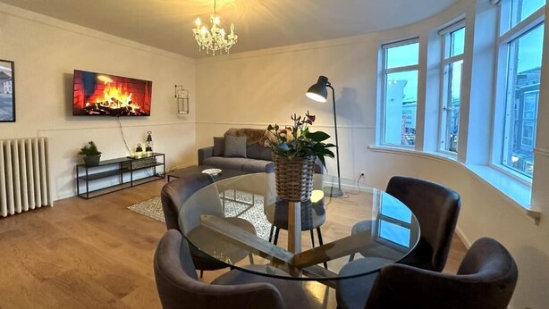 Een moderne woonkamer met een glazen eettafel, comfortabele zithoek, een open haard en grote ramen.