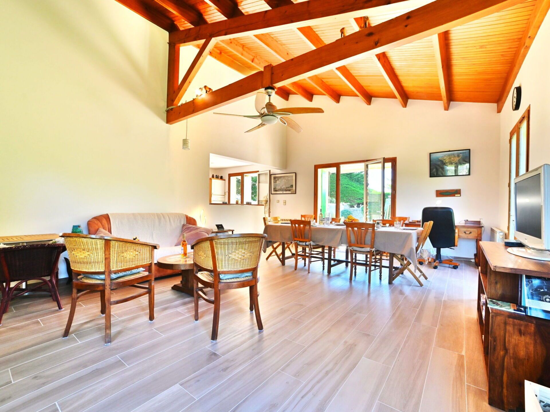 Una habitación amplia con vigas de madera, ventilador de techo, zona de comedor con mesa y sillas, y televisor.
