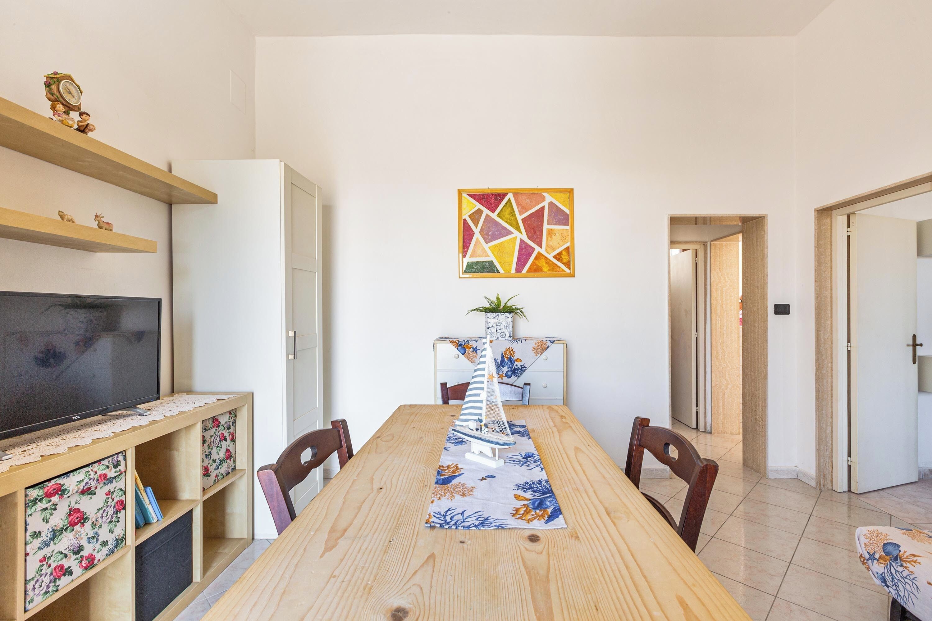Un comedor con una mesa de madera, sillas, un televisor sobre un soporte y un cuadro abstracto colorido en la pared.