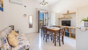 A living room with a sofa, a dining table with chairs, a TV, and a kitchen area with shelves. - 4188 Casa Rosy by Barbarhouse (Porto Cesareo)