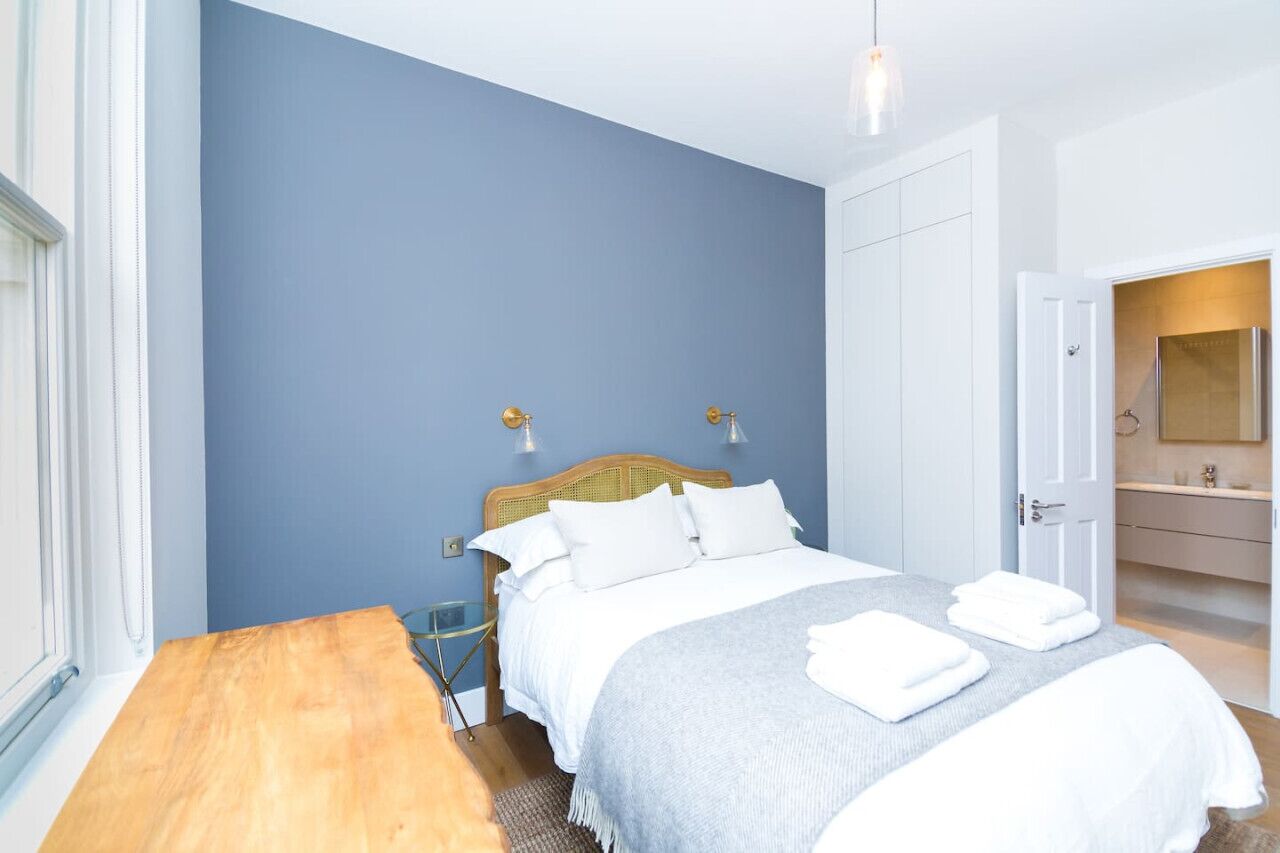 A bedroom with a bed, bedside tables, a wooden nightstand, a mirror, and a bathroom with a sink and mirror.