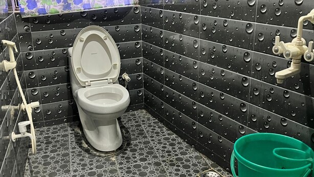 A bathroom with a toilet, a green bucket, and a shower area. - Thakur Ji P. Guest House By GRB (Varanasi)