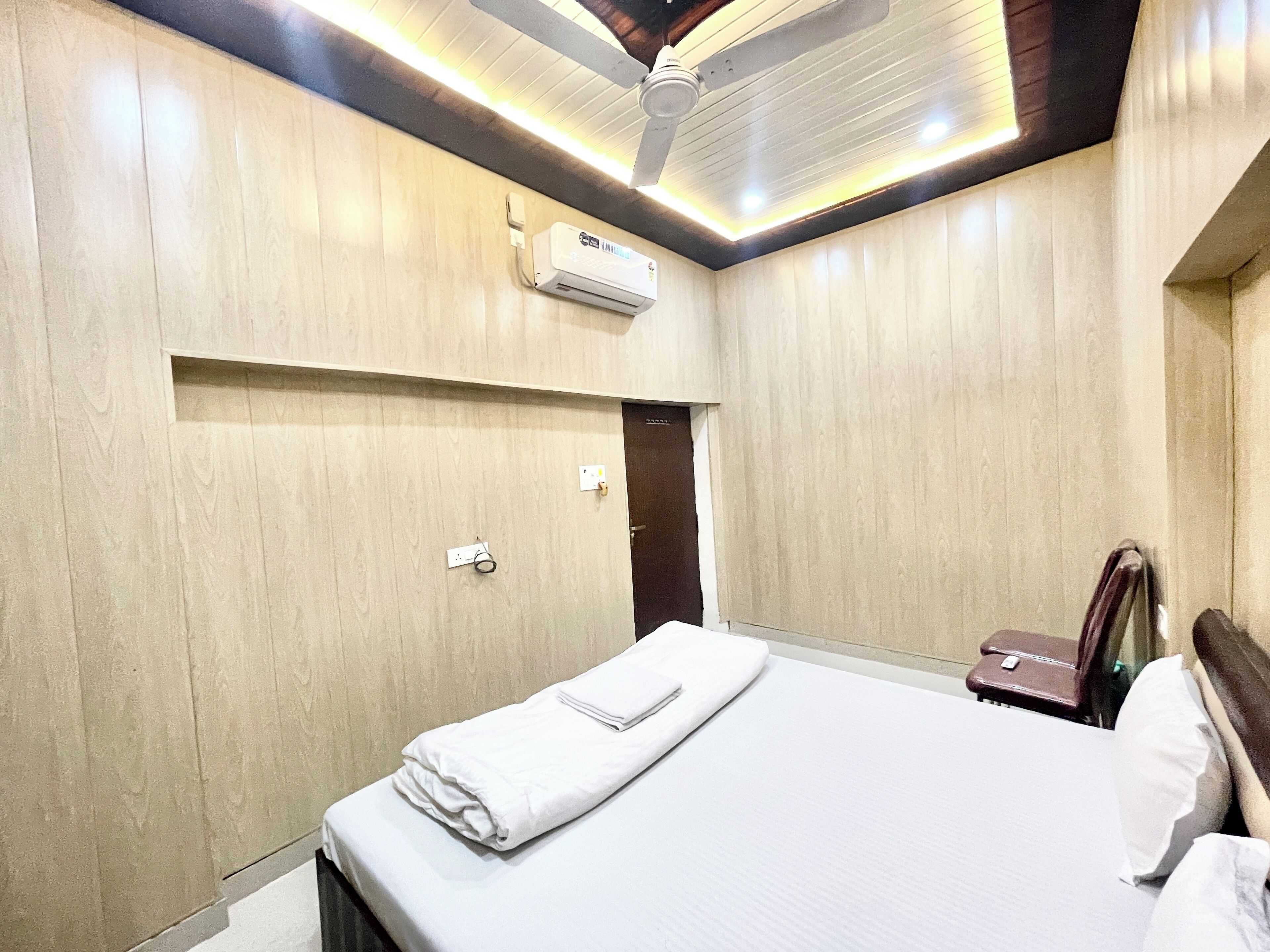 A hotel room with a bed, a ceiling fan, an air conditioning unit, and a small bedside table.