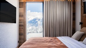 Een slaapkamer met een groot bed, houten wanden, een schuifdeur naar een balkon en uitzicht op besneeuwde bergen.