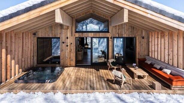 Een houten hut met een jacuzzi, buiten zitplaatsen en een groot raam met uitzicht op een besneeuwd landschap.