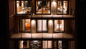 Een meerlaagse houten hut met een balkon, verlichting van binnen en een stenen muur.