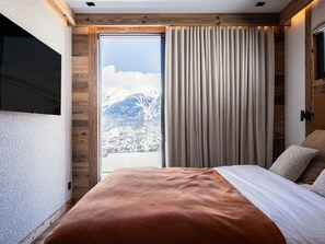 A bedroom with a large bed, wooden walls, a sliding door to a balcony, and a view of snow-covered mountains.