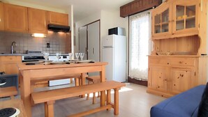 A compact kitchen with wooden cabinets, a dining table with benches, and a refrigerator. - Renovated 2-bedroom apartment in Samoëns, pets allowed (Samoëns)