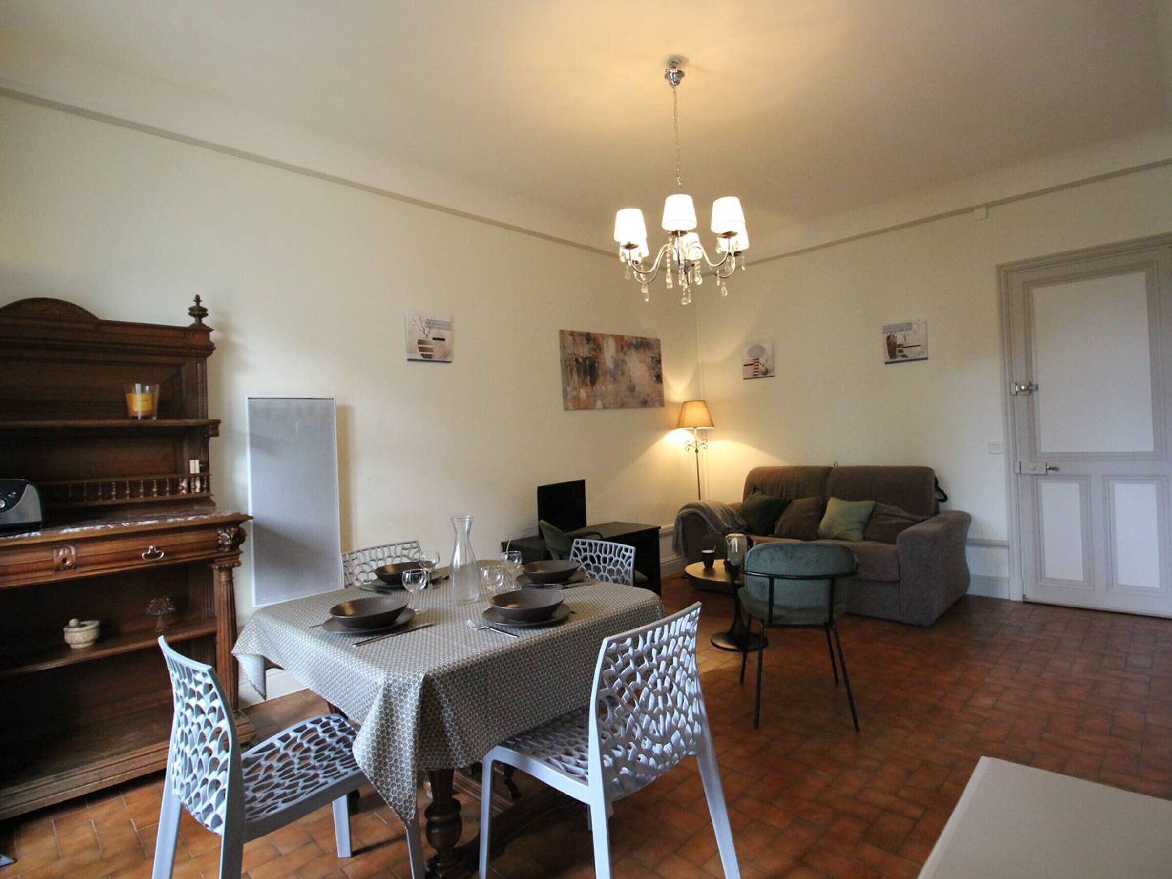 Un coin repas avec une table dressée pour quatre personnes, entourée de chaises à motifs. Un lustre est suspendu au plafond, et un canapé est visible au fond.