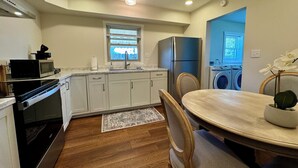 A compact kitchen with white cabinets, a built-in oven, microwave, and refrigerator. There's a window with blinds, a washing machine, and a dining area with a table and chairs. - Campus Den on Poplar St. (Marion)