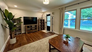 A living room with a wooden entertainment center, a flat-screen TV, a dining table, and a large window with a view of the outdoors. - Campus Den on Poplar St. (Marion)