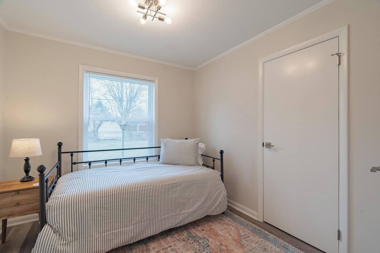 Een slaapkamer met een bed, nachtkastje, lamp, raam met zonwering en een deur.