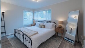 Ein Schlafzimmer mit Bett, Nachttischen, einer Stehlampe, einem Spiegel und einem Fenster mit Jalousien.
