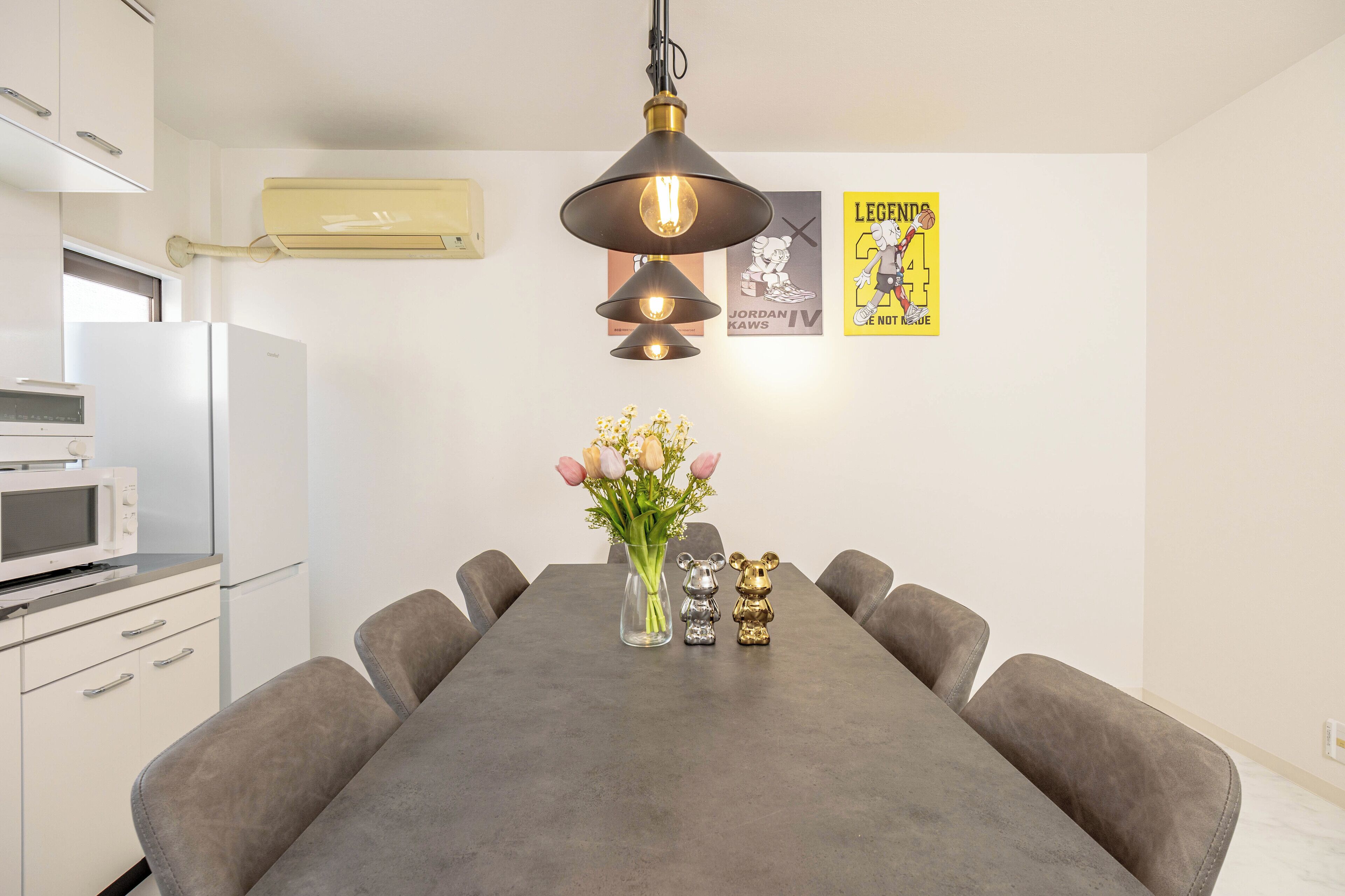 A dining area with a long table, chairs, a vase with flowers, a lamp, and a poster on the wall.