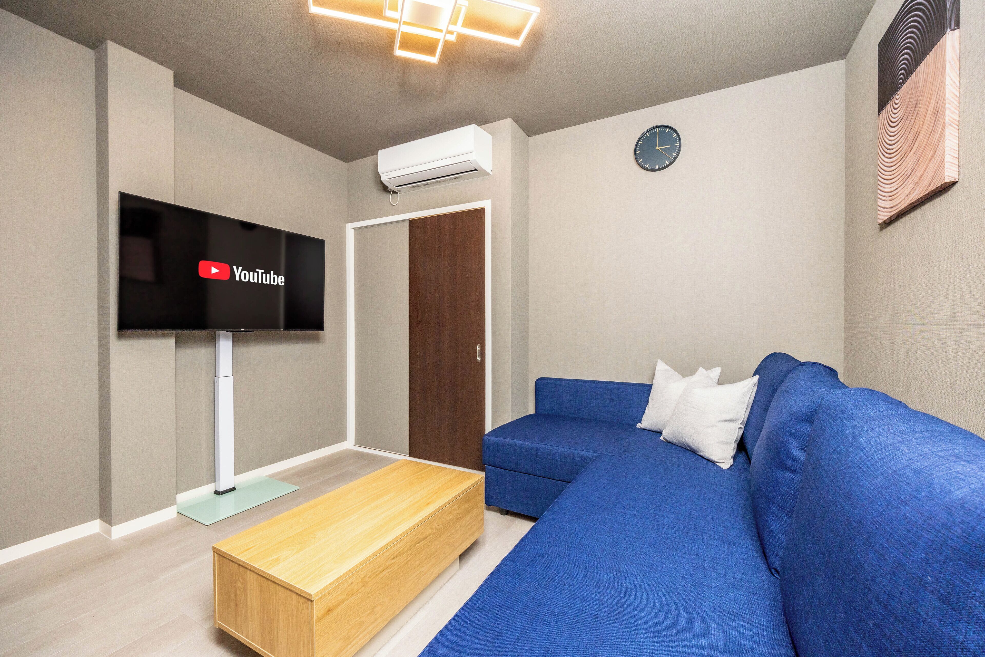 A modern living room with a blue sofa, a wooden coffee table, a flat-screen TV mounted on the wall, and a clock on the wall.