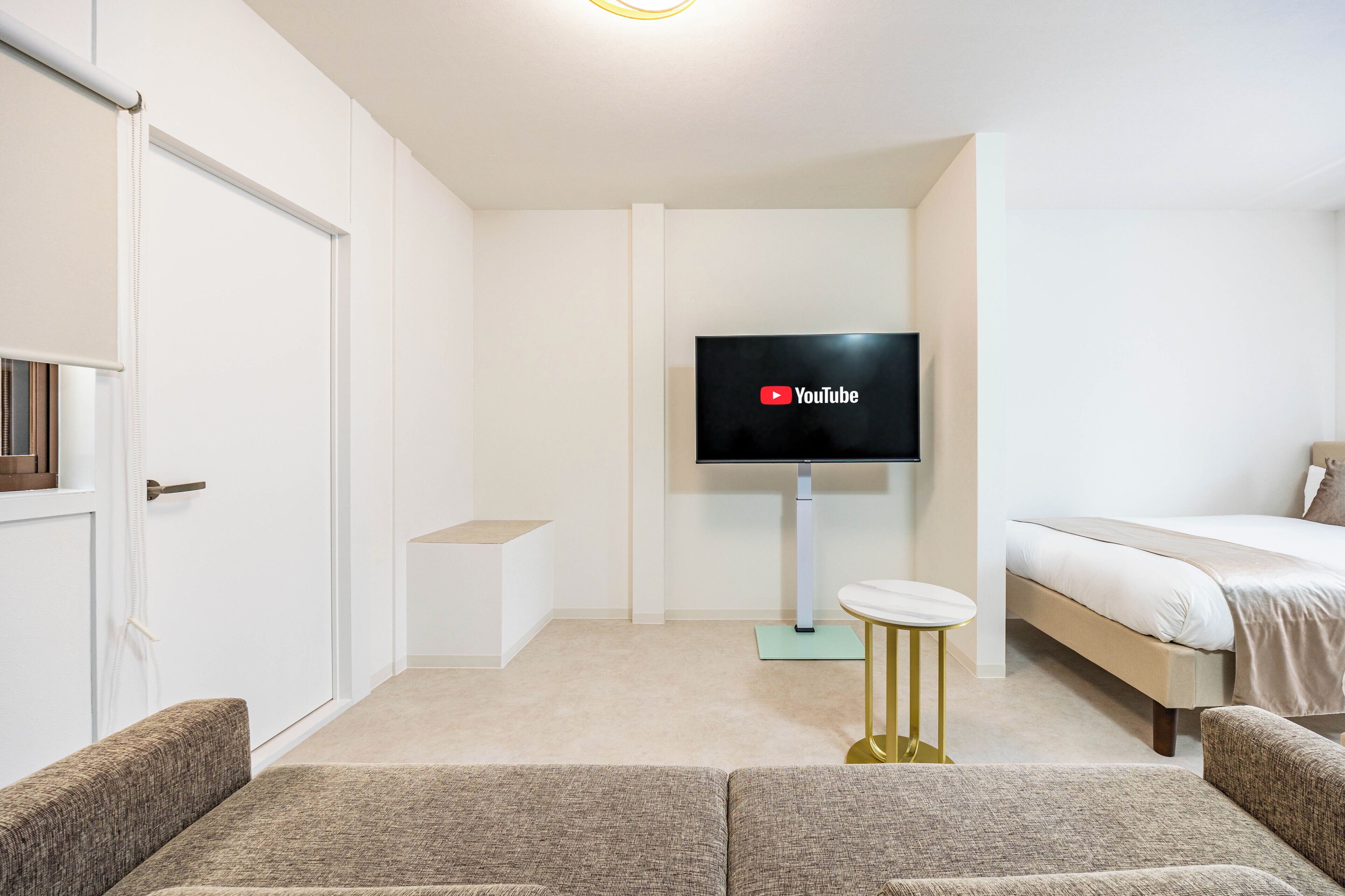 Una habitación de hotel moderna con un televisor de pantalla plana montado en la pared, una cama con colchón blanco y ropa de cama color beige, una mesita redonda con base dorada y un sofá color beige.