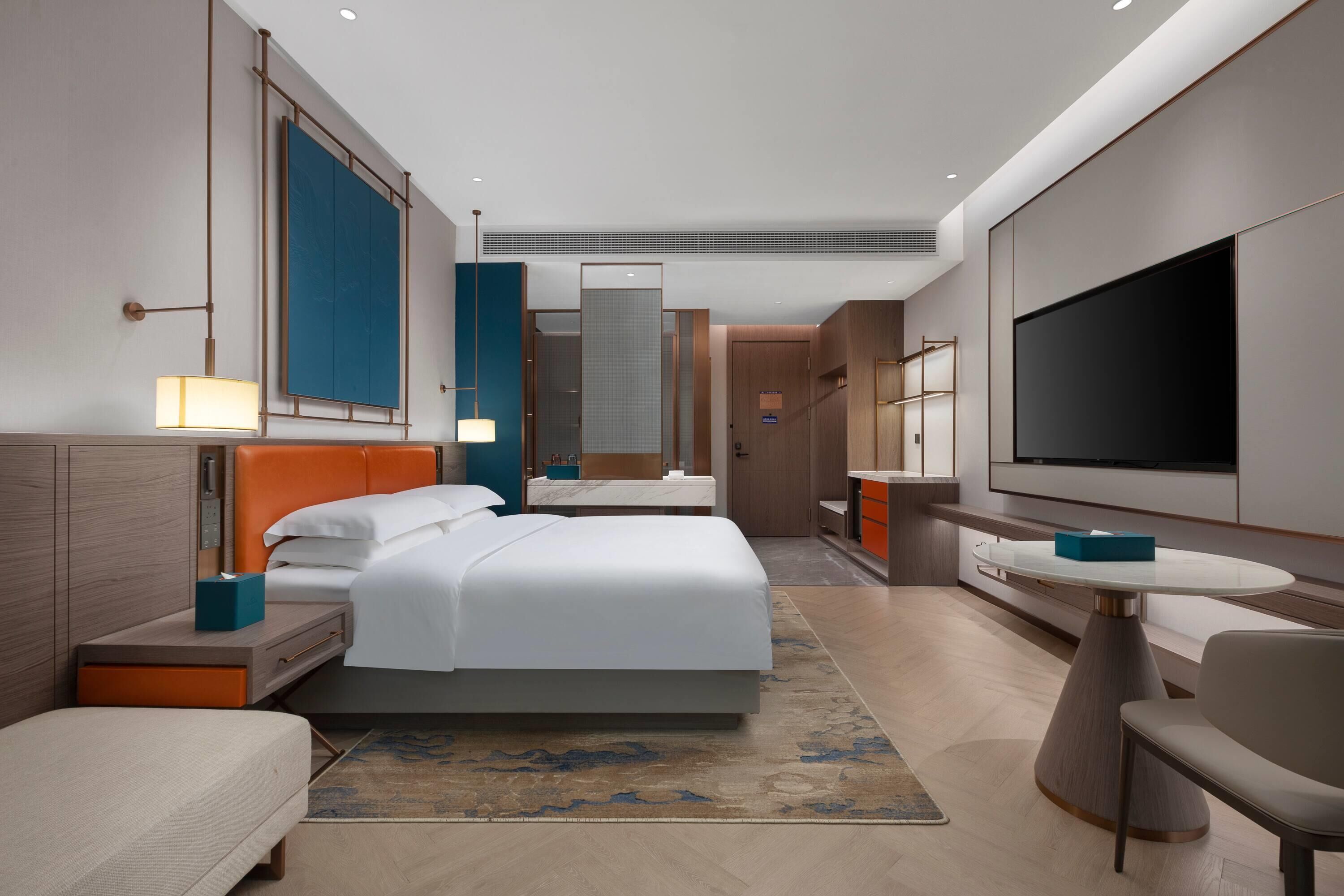 A modern hotel room with a large bed, a TV, a desk, and a seating area.