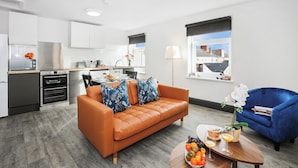 A modern kitchen with built-in appliances, a dining area with a table and chairs, and a living room with a sofa and a blue armchair. - TYME Suites on Humber (Cleethorpes)