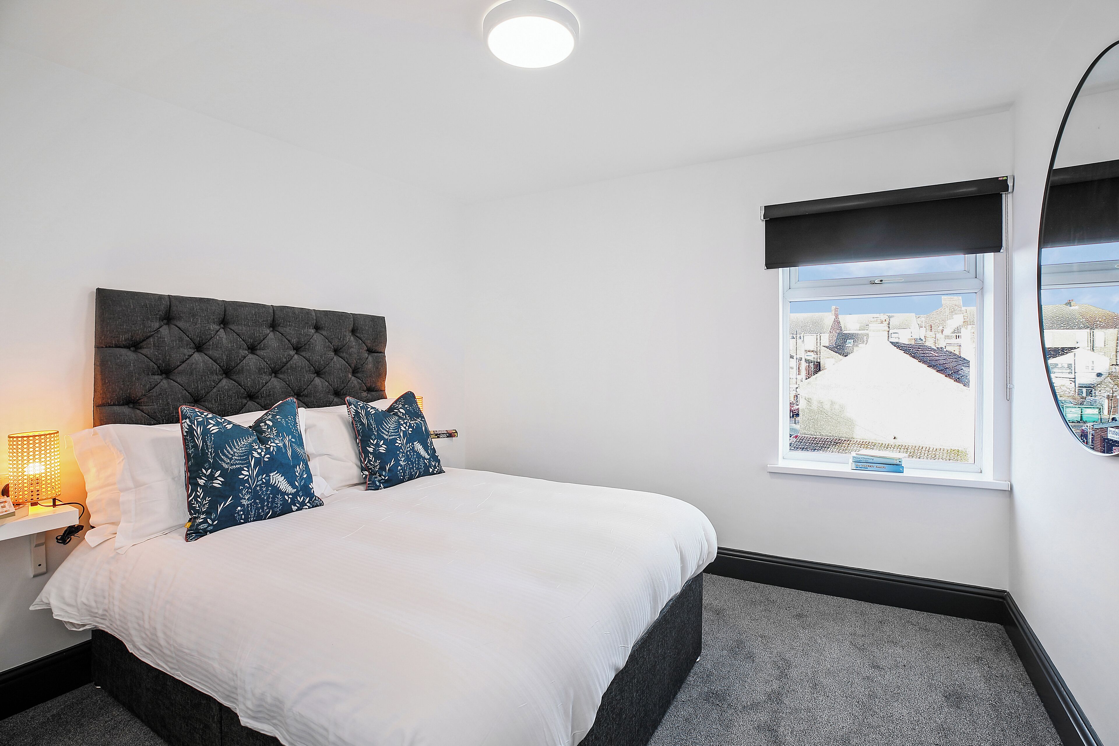 A neatly made bed with white and blue patterned pillows, a dark tufted headboard, and a window with a view of buildings.
