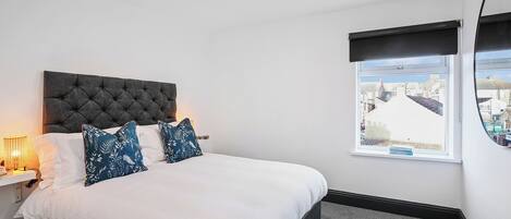 A neatly made bed with white and blue patterned pillows, a dark tufted headboard, and a window with a view of buildings.