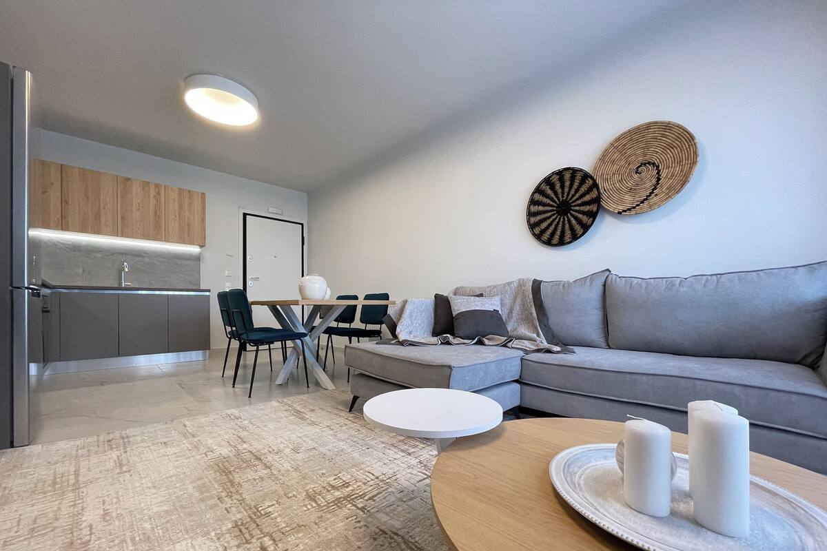 Una sala de estar moderna con un sofá gris, una mesa de centro redonda y un comedor con una mesa y sillas blancas.