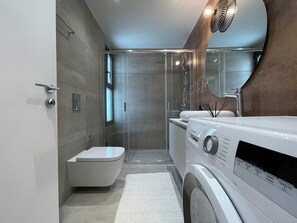 A modern bathroom with a Bosch washing machine, a toilet, and a glass-enclosed shower. - Urban Oasis in Ellinikon (Elliniko-Argyroupoli)