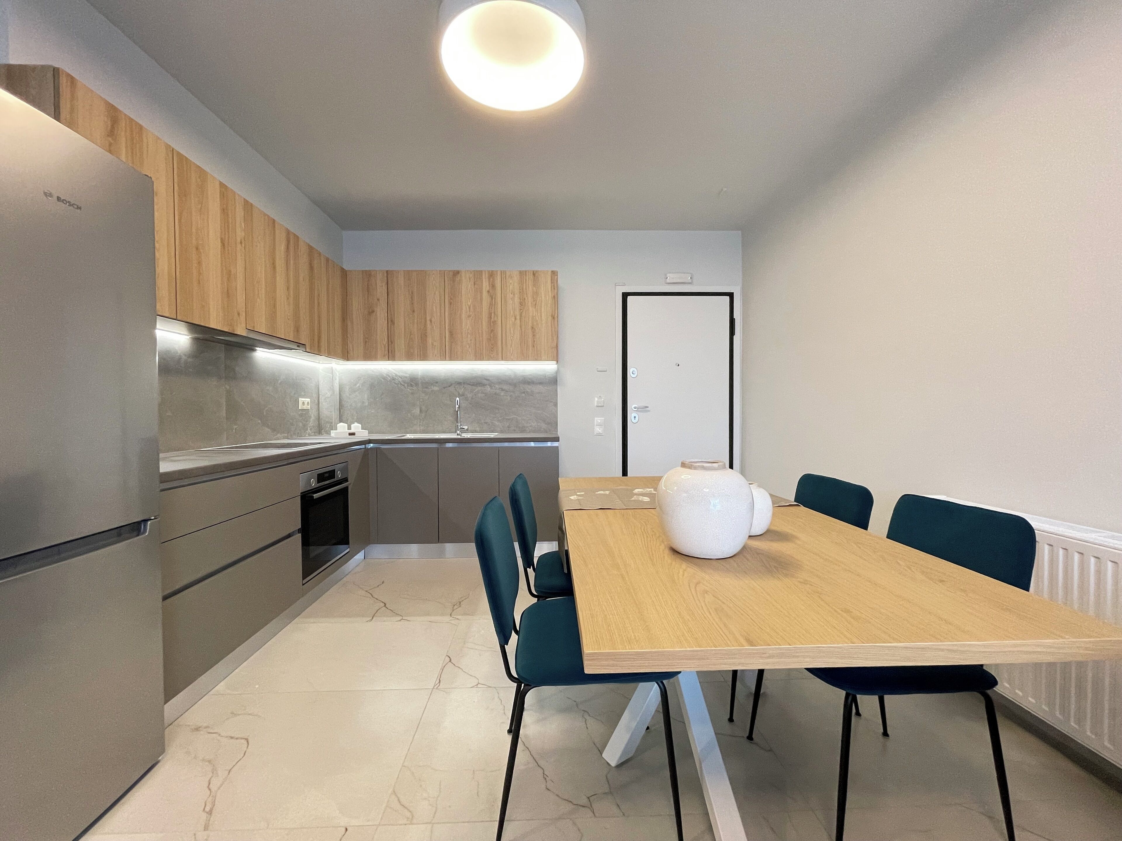 Una cocina moderna con horno empotrado, un lavamanos y armarios de madera. Hay una mesa de comedor con sillas y un florero blanco sobre ella.