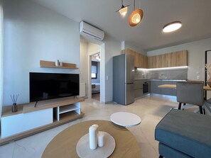 A modern living room with a flat-screen TV, a sofa, a coffee table, and a kitchen area with wooden cabinets and a built-in oven. - Urban Oasis in Ellinikon (Elliniko-Argyroupoli)