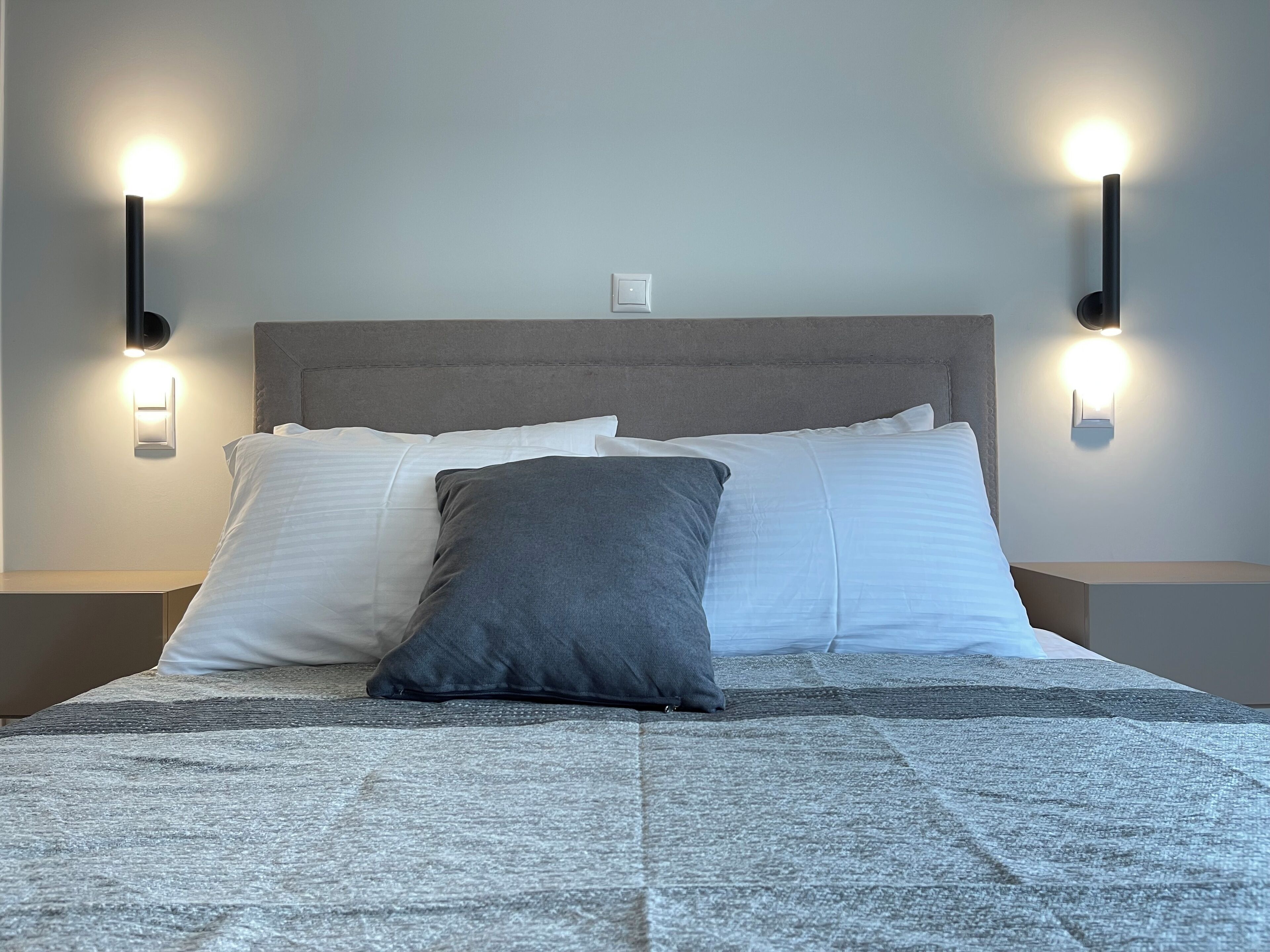 A neatly made bed with white and gray bedding, two wall-mounted nightstands, and two wall-mounted light fixtures.