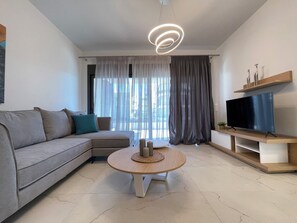 Un salon moderne avec un canapé, une table basse ronde, un téléviseur à écran plat posé sur un meuble en bois et un luminaire suspendu au plafond.
