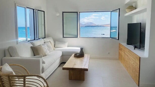 Un soggiorno moderno con un divano bianco, un tavolino in legno e una TV a parete. Ampie finestre offrono vista sul mare e su una montagna all'orizzonte.