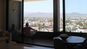 Una sala de estar moderna con un ventanal grande que ofrece vistas a un paisaje urbano y montañas. Hay un comedor con mesa y sillas.