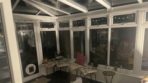 A conservatory with a glass door, a small table with a chair, a red cushion, and a water bottle on a windowsill. - Rochester Retreat - Cozy 3 Bedroom Vacation Home (Rochester)