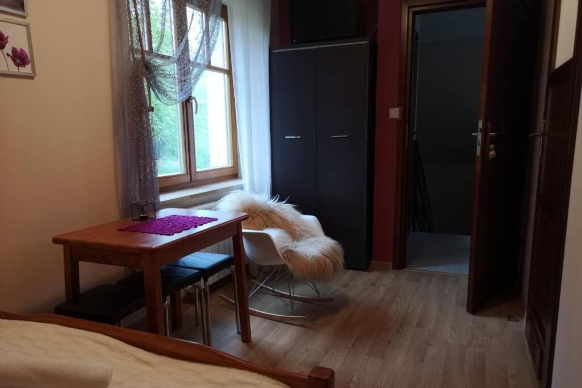 A bedroom with a bed, a wooden desk, a rocking chair, a wardrobe, and a window with curtains.