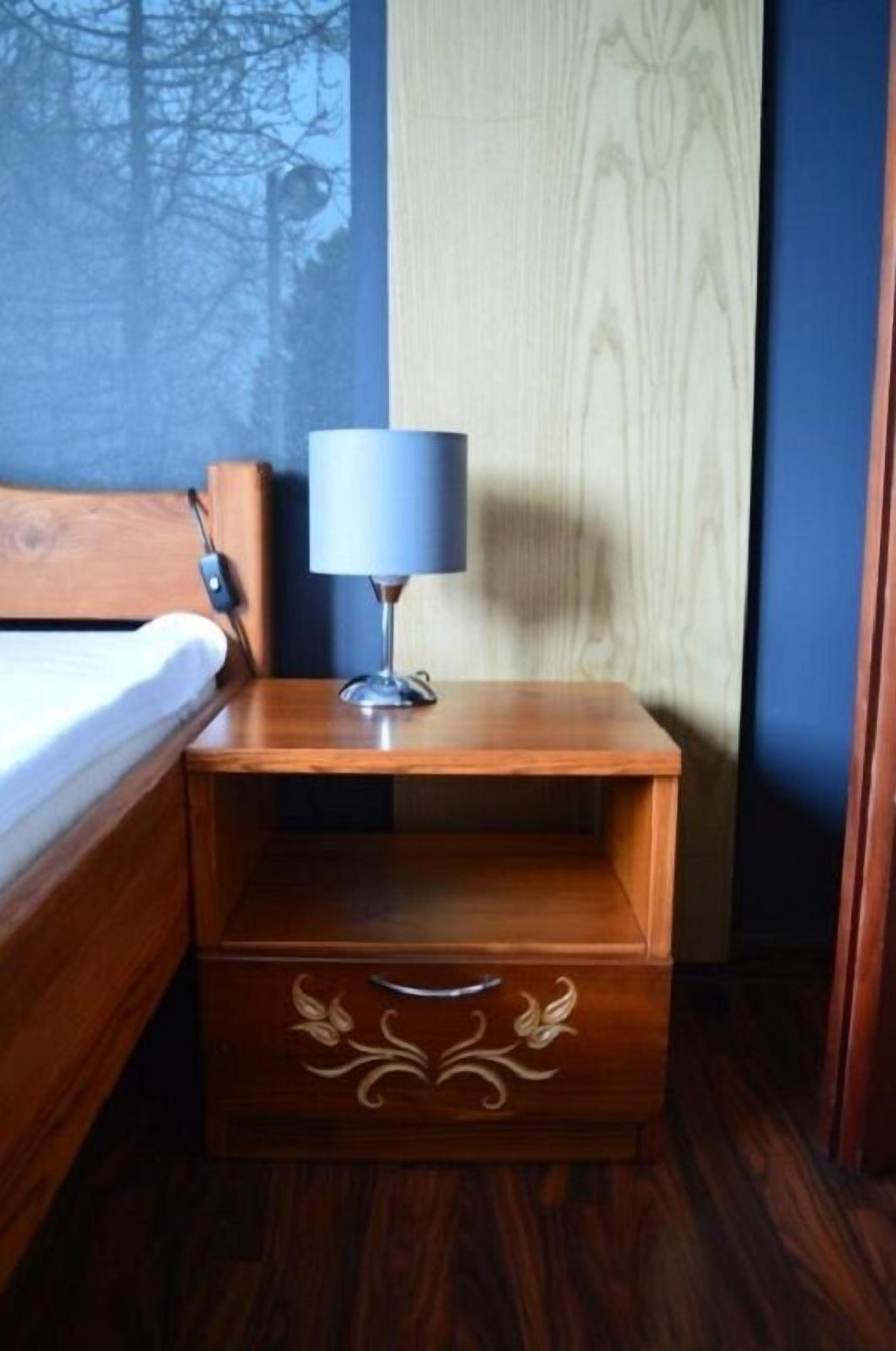A wooden bedside table with a lamp, a drawer with floral carvings, and a bed with a white sheet.