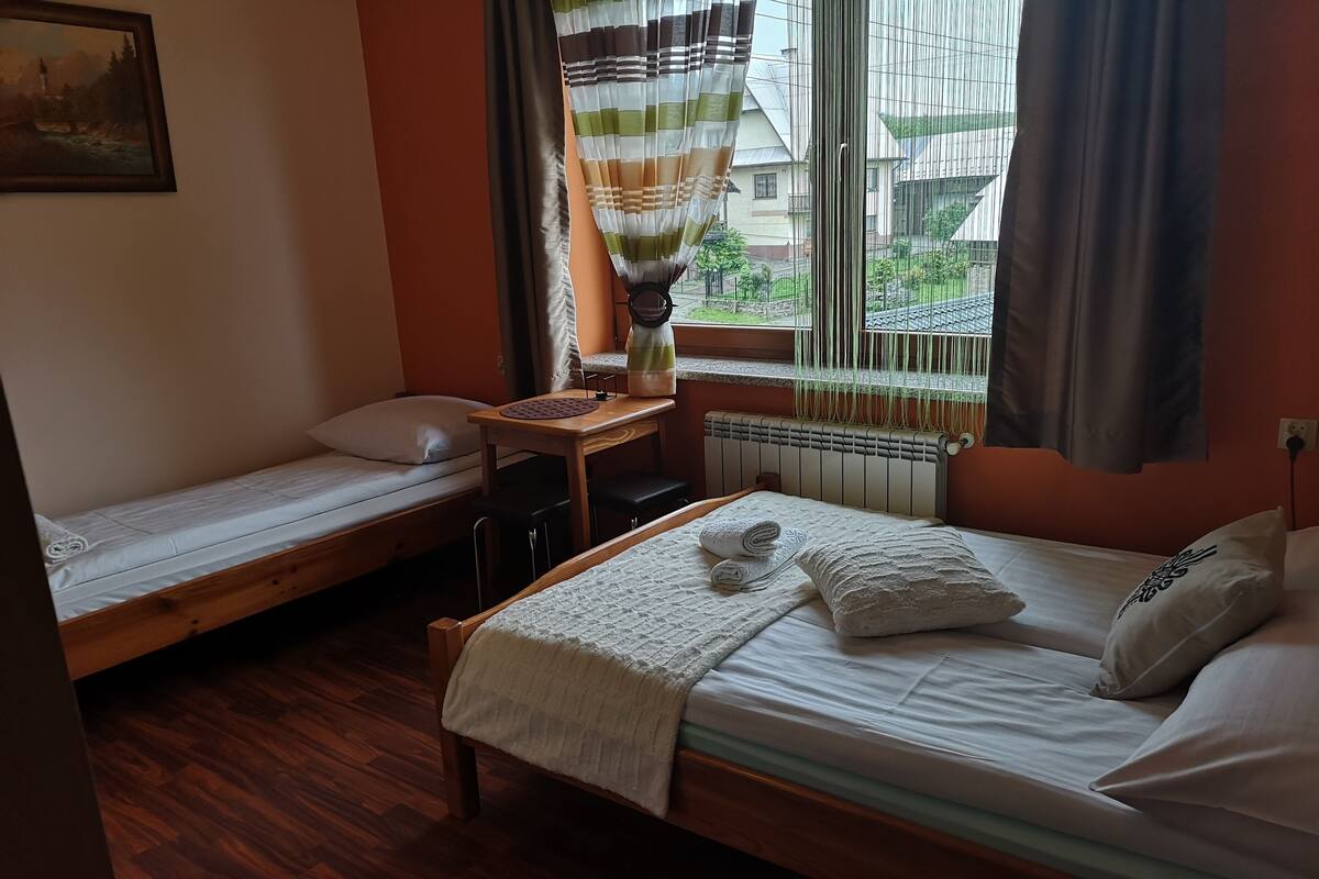 A small, cozy room with two single beds, a wooden table, a radiator, and a window with curtains.
