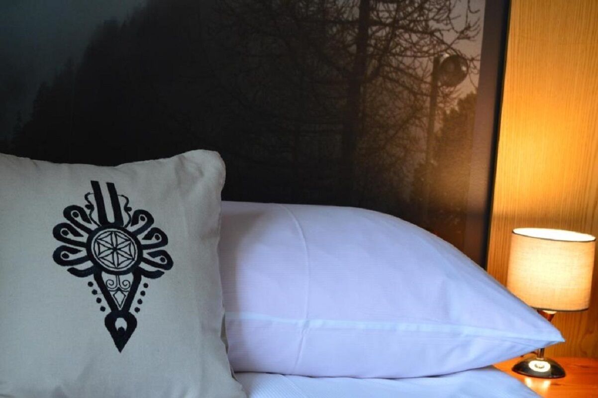 A bed with a white pillow featuring a geometric design, a white pillow, and a bedside lamp on a wooden nightstand.