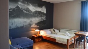 Une chambre à coucher comprenant un lit, une table de chevet avec une lampe, une chaise, une petite table et une grande fresque murale représentant un paysage de montagne.