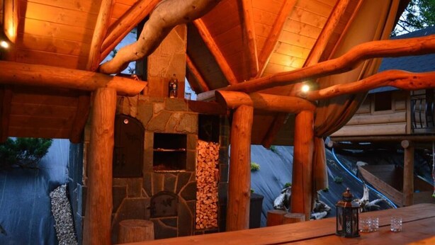Une structure rustique en bois, dotée d’une cheminée en pierre, de poutres apparentes et d’une table sur laquelle se trouvent des verres et une lanterne.
