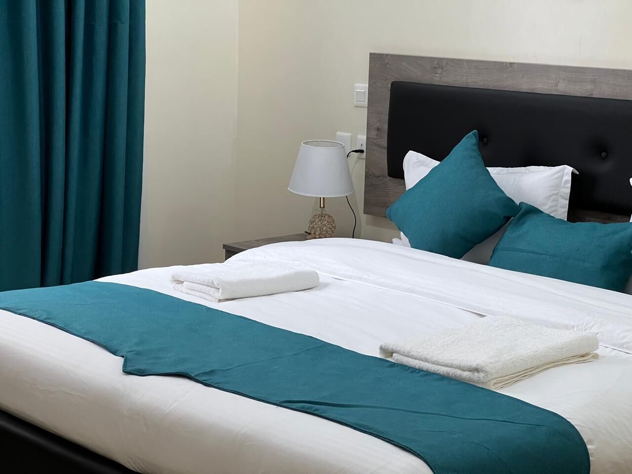 Una cama bien hecha con ropa de cama azul verdoso y blanca, una lámpara de noche y cortinas azul verdosas.
