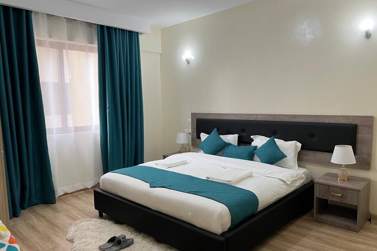 Une chambre moderne avec un grand lit, une literie aux couleurs turquoise et blanche, un tapis moelleux et une table de chevet avec une lampe.