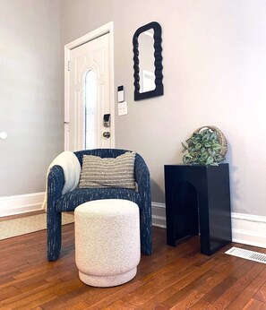 Um quarto com porta, uma cadeira, um pequeno banquinho, uma mesinha de cabeceira preta com uma planta e um espelho fixado na parede.