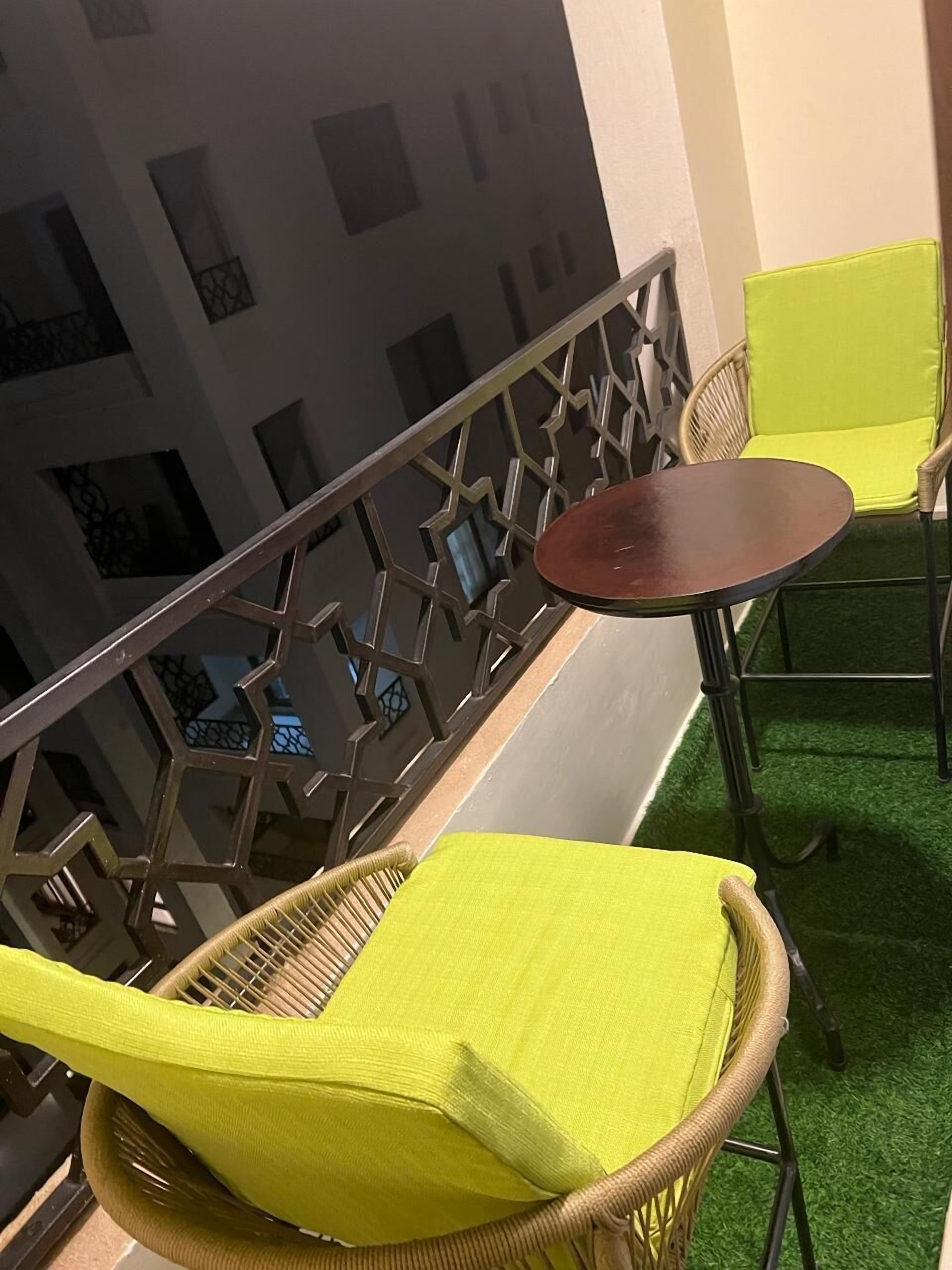 Un balcón con sillones de mimbre y una pequeña mesa redonda, con barandilla de diseño geométrico y piso cubierto de césped artificial verde.